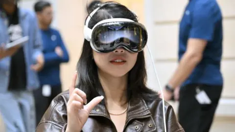 Getty Images A woman using the Vision Pro headset inside an Apple store.  It looks like a pair of sleek, shiny, curved, futuristic glasses strapped to her face.  You could almost say it looks nice.